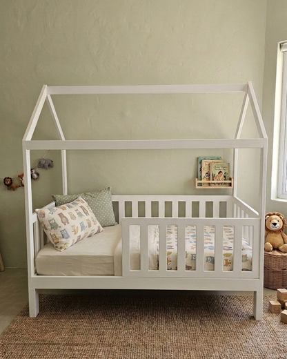 White crib with cushions and a teddy bear in a room with a green wall and wooden floor.