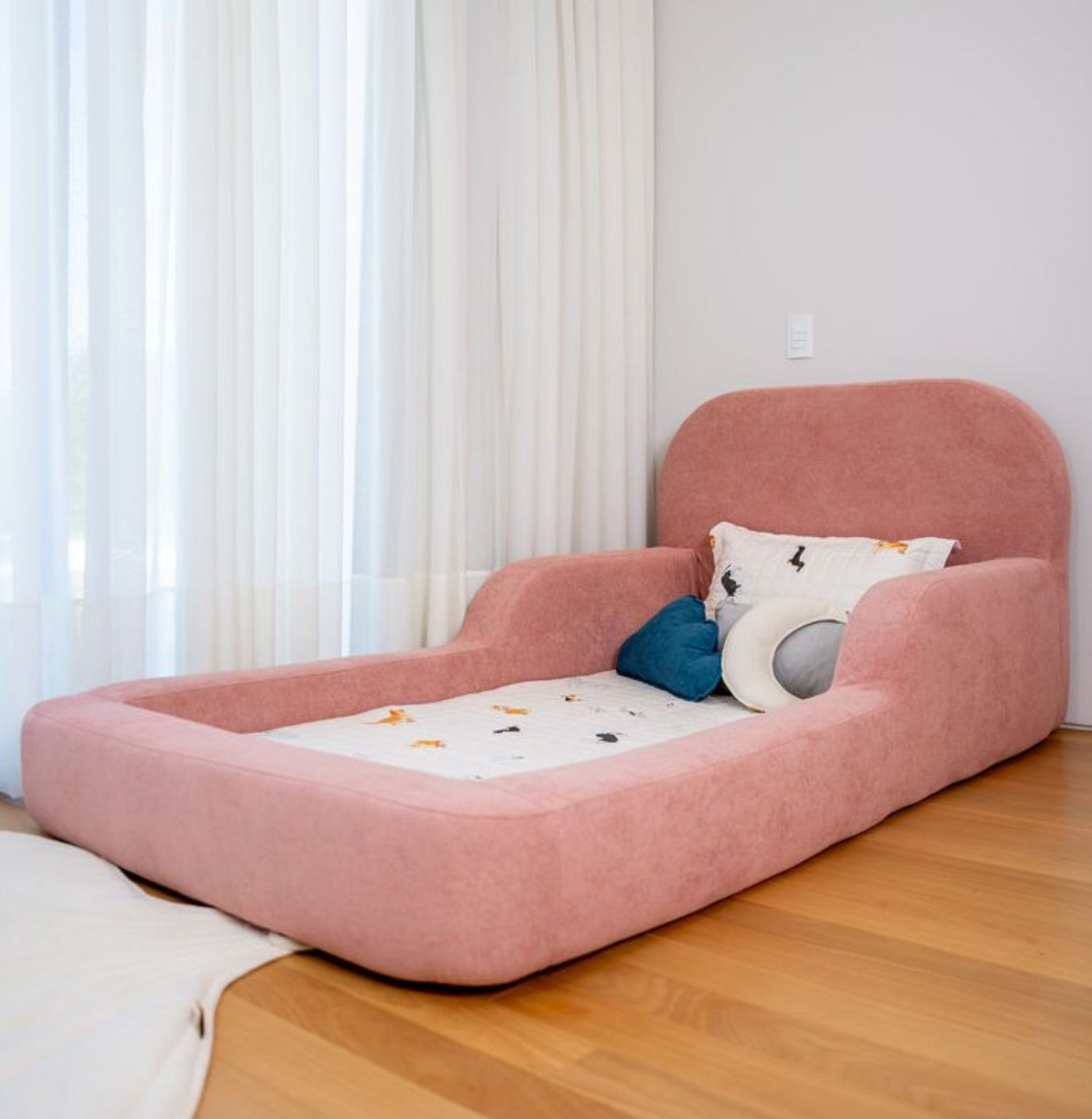 Pink children's bed with cushions in a room with white curtains and wooden floor.